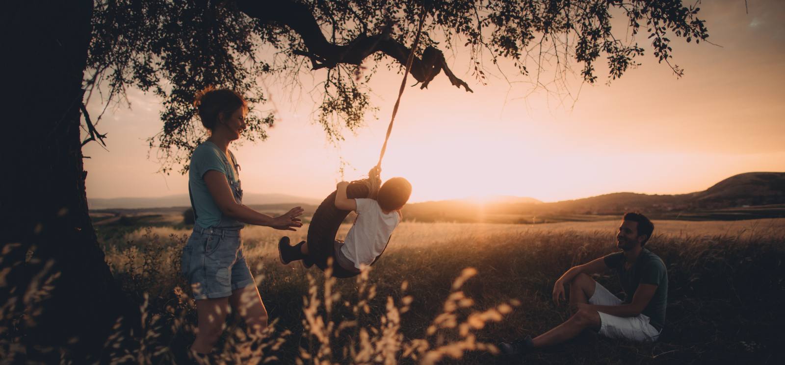 Mom pushing child on tire swing while the sun sets.